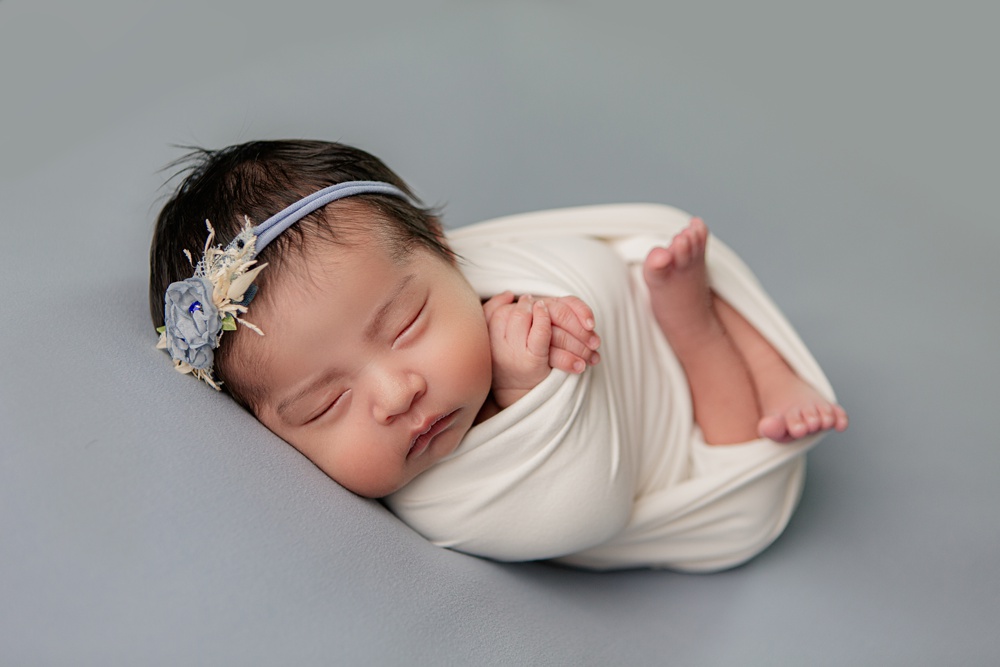 Newborn girl wrapped in white with blue headband on blue backdrop
